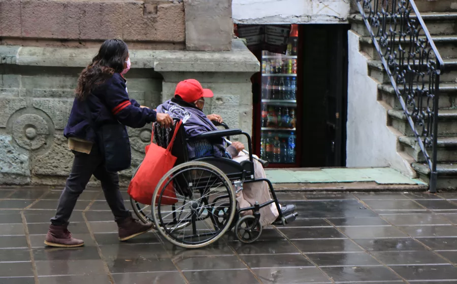 Usuaria en silla de ruedas guiada por una mujer sobre la acera mojada.