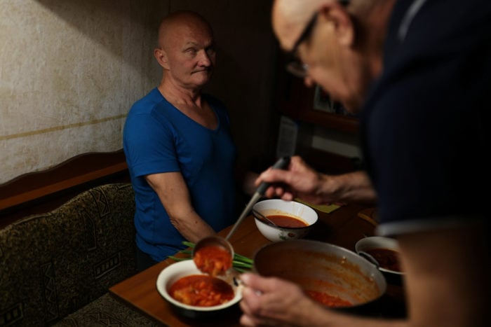 Viktor Solovyanenko sentado a oscuras en una mesa, otro compañero busca el cucharón de sopa para servirse en un tazón.