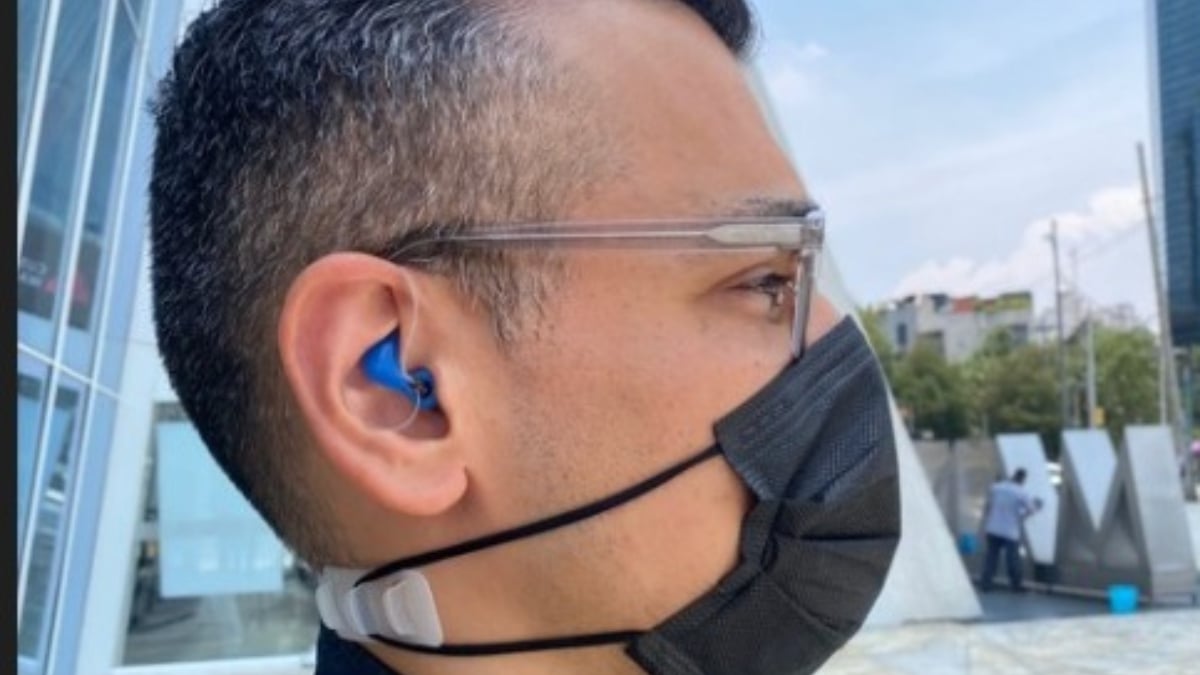 Fotografía de un hombre de perfil, que usa cubrabocas negro, lentes transparentes y un audífono azul de un centímetro o menos de tamaño