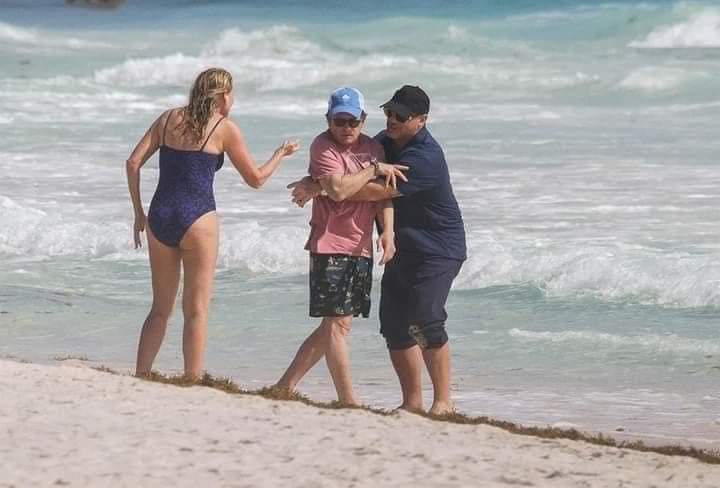 Michael J. Fox cerca de las olas del mar ayudado por un cuidador para continuar de pie.