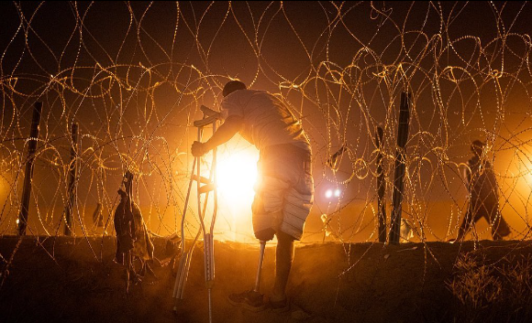 Migrante usuario de prótesis en la pierna izquierda, deja de lado sus muletas para cruzar una cerca de aluminio con alambre de púas