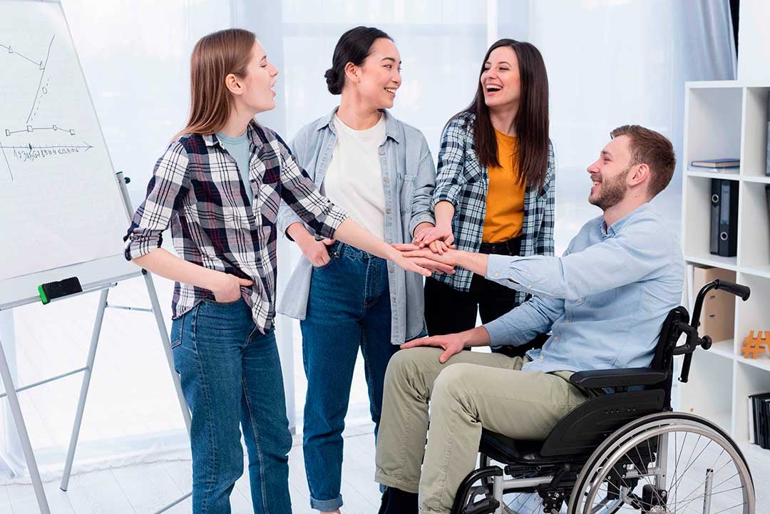 Cuatro personas uniendo sus manos dentro de una oficina, uno de ellos es un usuario de silla de ruedas