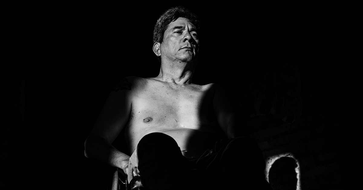 Fotografía en blanco y negro de un hombre en silla de ruedas, en donde se alcanza a ver parte de su pecho y su cara, tiene una mirada desafiante pero parece esconderse en la sombra