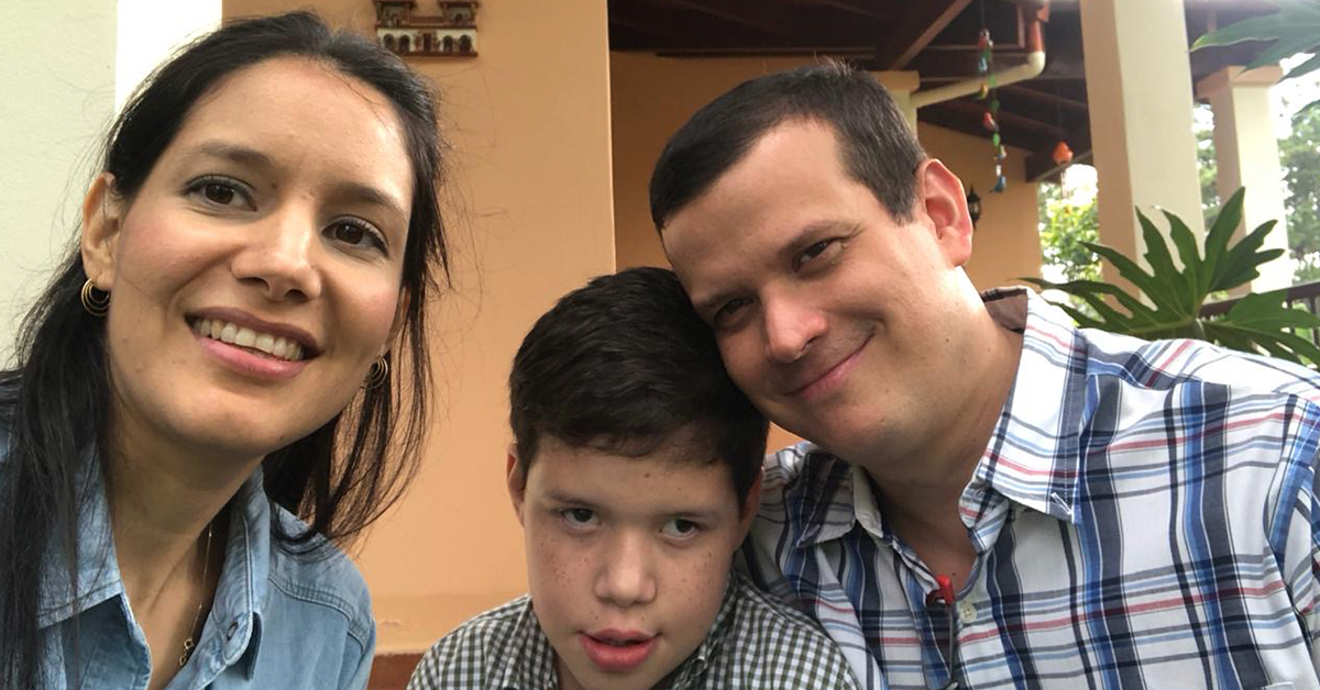 Fotografía de Di con sus papás, Karola y Dieter. En la fotografía los tres están sonriendo, su papá usa una camisa de rayas y su mamá una camisa de mesclilla azul, Di tiene una camisa de cuadritos, todos ven a la cámara.