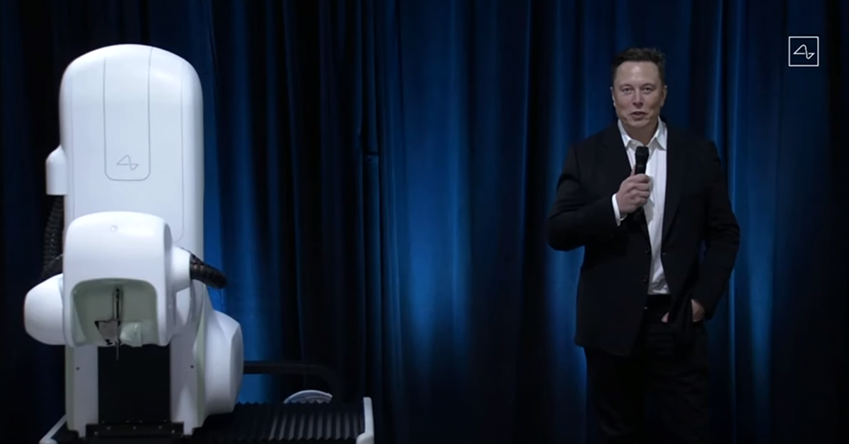 Fotografía de cuerpo completo de Elon Musk, en la fotografía Elon viste un traje negro con una camisa blanca y corbata negra, sostiene en su mano derecha un micrófono, a la derecha de Elon está una de las partes del neurolink, es una maquina grande color blanco. Detrás de ellos una cortinilla color azul.