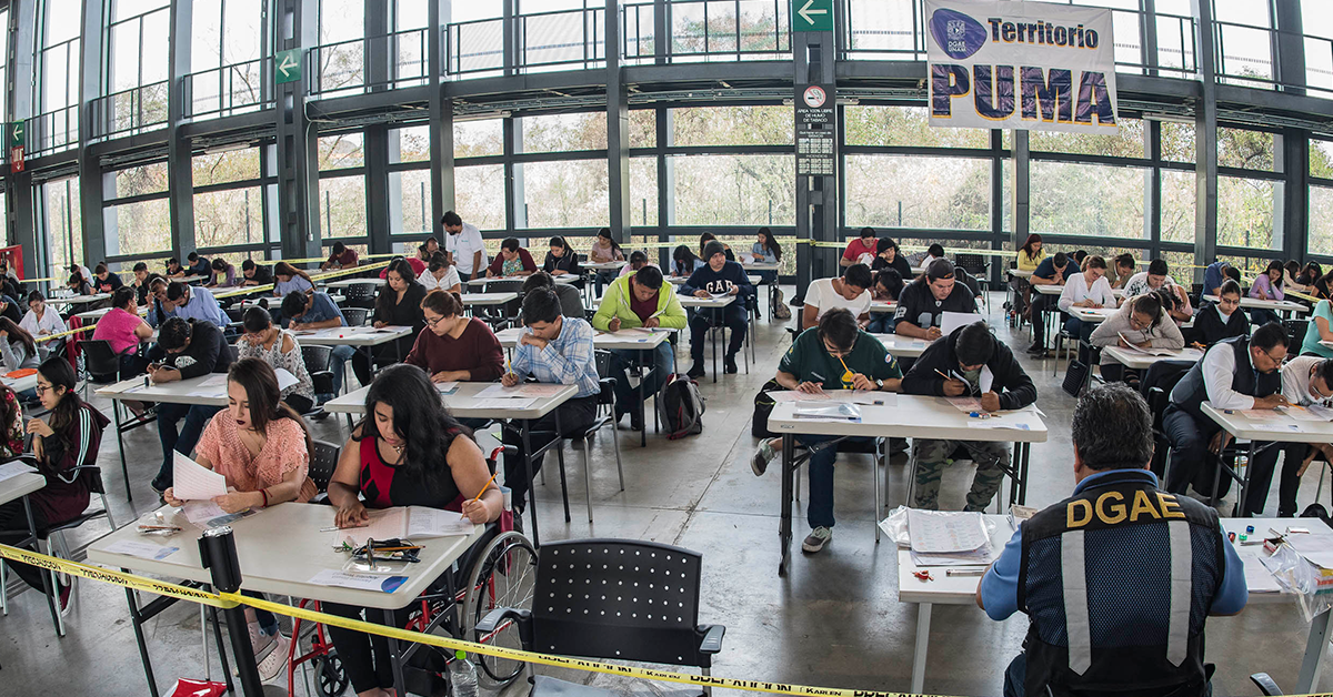 Fotografía de uno de los grupos de estudiantes presentando su examen para ingresar a la UNAM, al frente una mujer en silla de ruedas vestida de rojo, detras de ella una fila de 4 escritorios, cada uno con 2 estudiantes, todos viendo su hoja de examen con un lápiz en la mano.