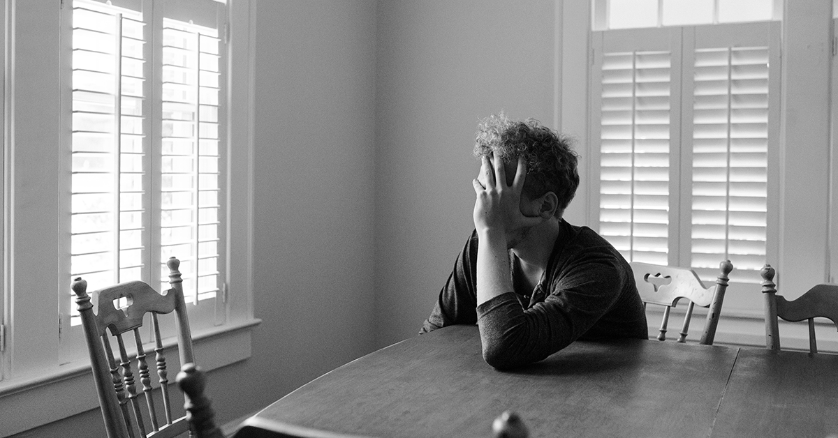 Fotografía en blanco y negro de un hombre sentado detrás de una mesa cubriendo su rostro con su mano.