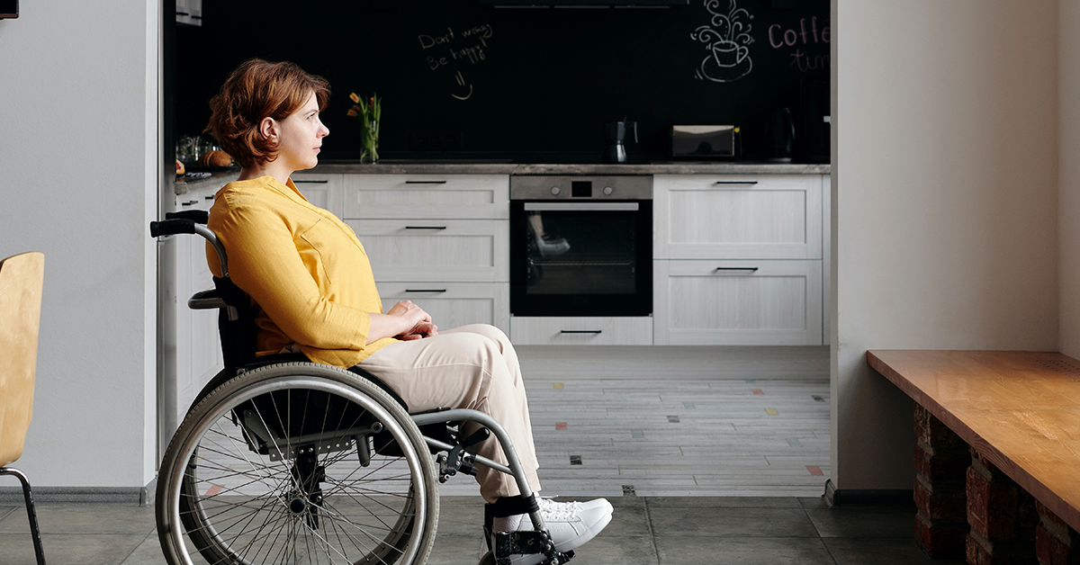 Fotografía de perfil de una mujer en silla de ruedas, detrás de ella su cocina y frente a ella su ventana.