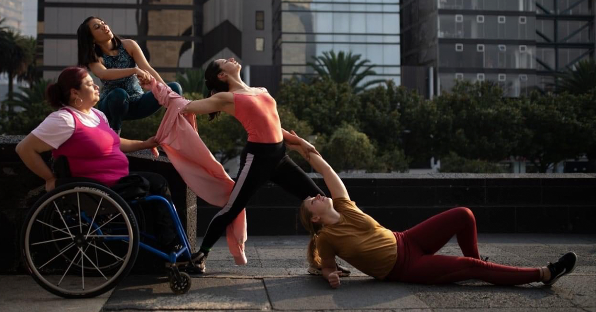 Fotografía de una coreografía. De izquierda a derecha, una mujer en silla de ruedas con una blusa rosa, junto a ella una mujer agachada agarrando un brazo de otra mujer frente a ellas que esta vestida con blusa rosa y un pantalón negro.