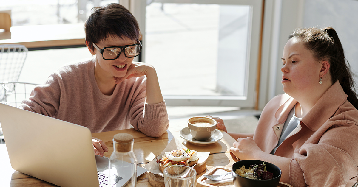 Fotografía de dos personas sentadas, a la izquierda una mujer con el cabello corto, lentes con el marco negro y una blusa rosa, a la derecha una mujer con sindrome de down con un saco color rosa. Sobre la mesa frente a ellas una computadora y platos de comida.