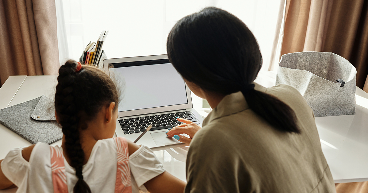 Fotografía de una mujer con su hija de espaldas frente a una computadora, la mujer tiene las manos en la computadora mientras la niña tiene un lápiz en la mano.