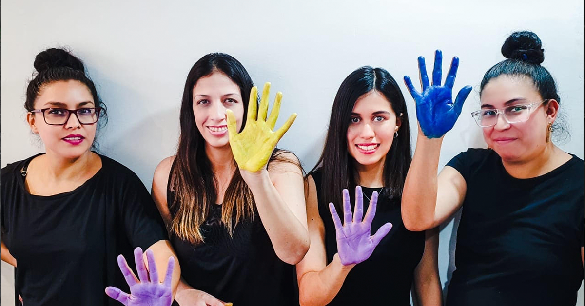 Fotografía de cuatro mujeres, todas tienen una mano pintada y levantada, de izquierda a derecha, la primera tiene pintura morada, la segunda amarilla, la tercera morada y la cuarta azul, todas tienen una playera negra.