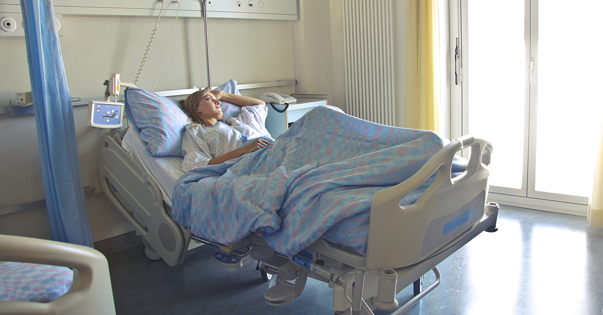 Fotografía de una mujer recostada en una cama de hospital, esta cubierta hasta la cintura con una cobija azul, tiene un brazo en el pecho y el otro en la cabeza, esta viendo hacia la izquierda.