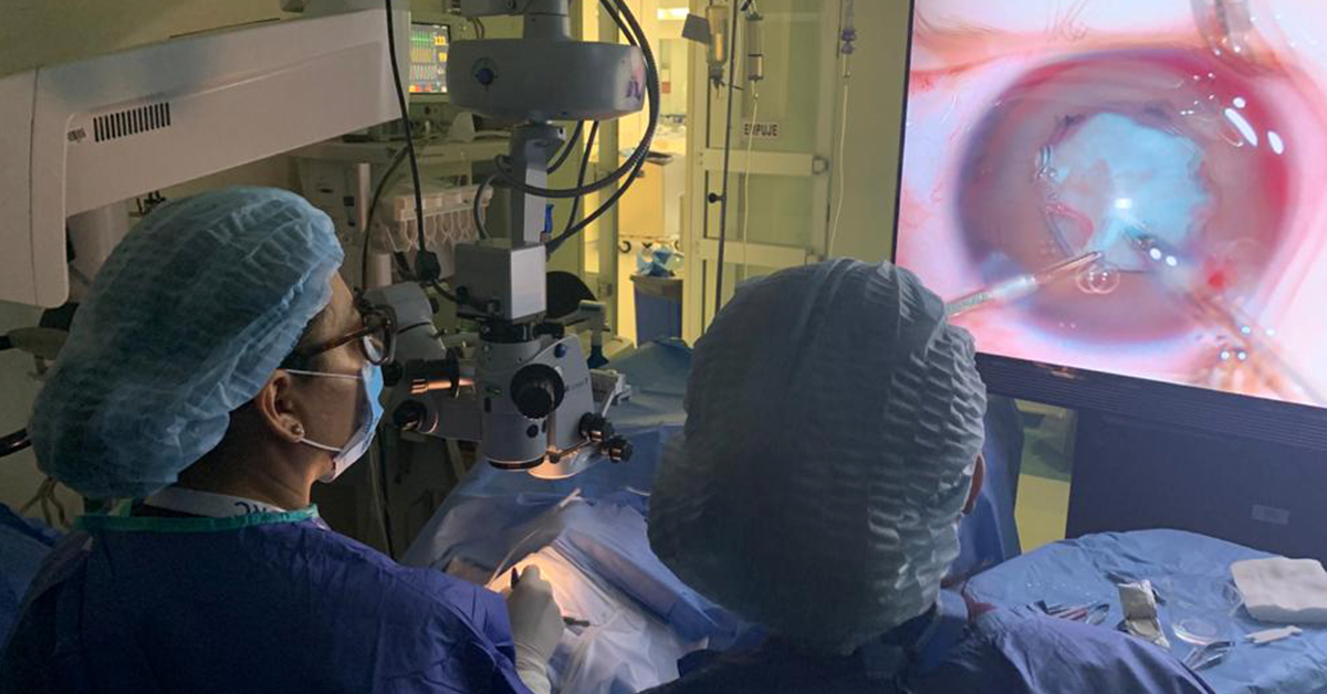 Fotografía de dos doctores de espaldas con una bata y gorro color azul, ven una pantalla donde se ve un ojo.
