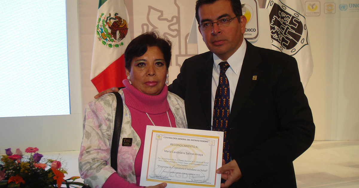 Fotografía de María Candelaria con una sudadera rosa sosteniendo un diploma con su mano derecha, junto a ella un hombre en traje negro y detrás de ellos la bandera de México.