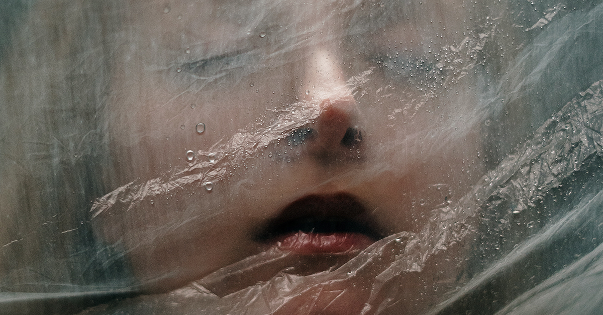 Fotografía del rostro de una mujer con los ojos cerrados detrás de una cortina de plástico transparente.