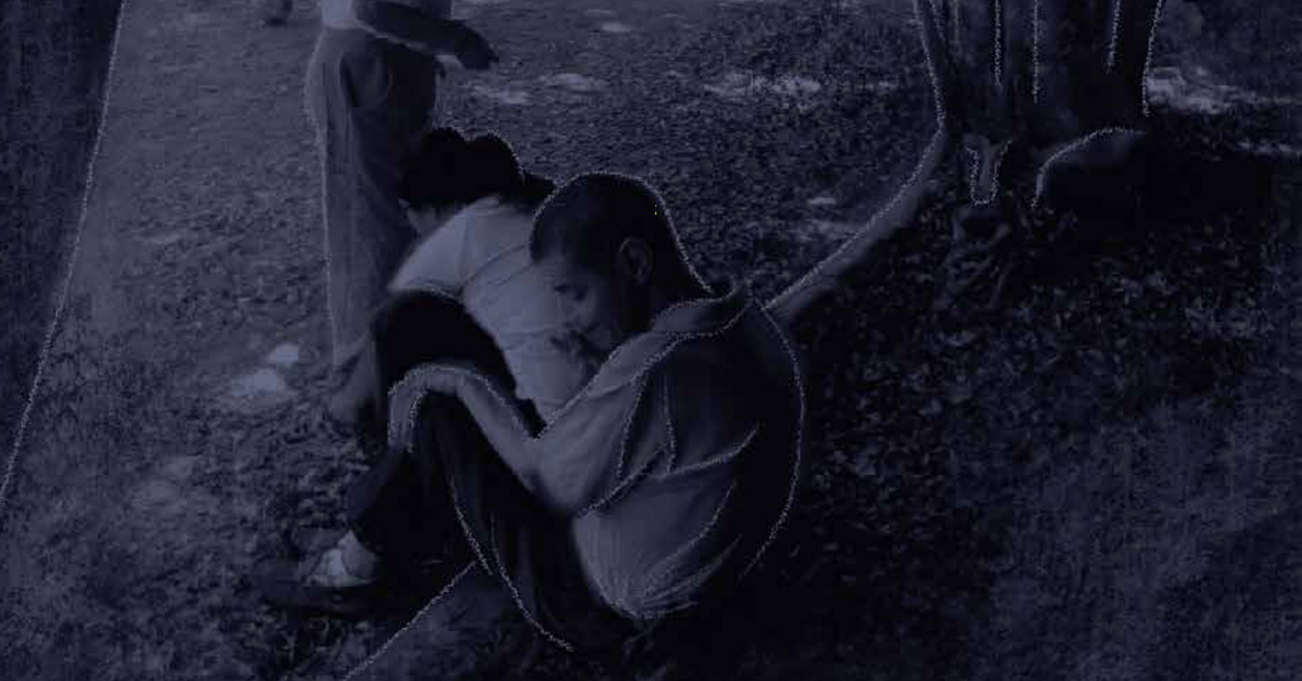 Fotografía en blanco y negro de un hombre y una mujer sentados sobre el tronco de un árbol, ambos ven al suelo.