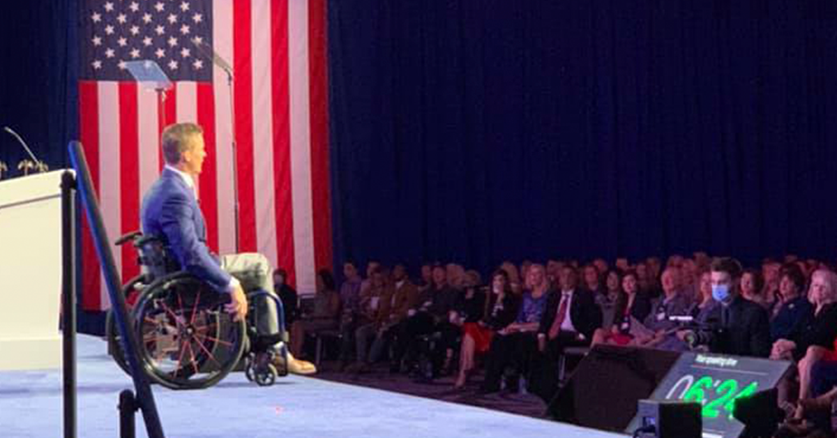 Fotografía de Madison sentado en su silla de ruedas, detrás de él la bandera de Estados Unidos y frente a él un grupo de personas viéndolo.