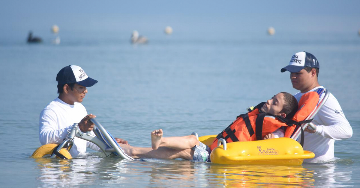 Fotografía de tres personas dentro del mar, dos de ellas vestidas con una playera blanca y una gorra azul ayudan a un hombre con discapacidad a flotar dentro del mar.