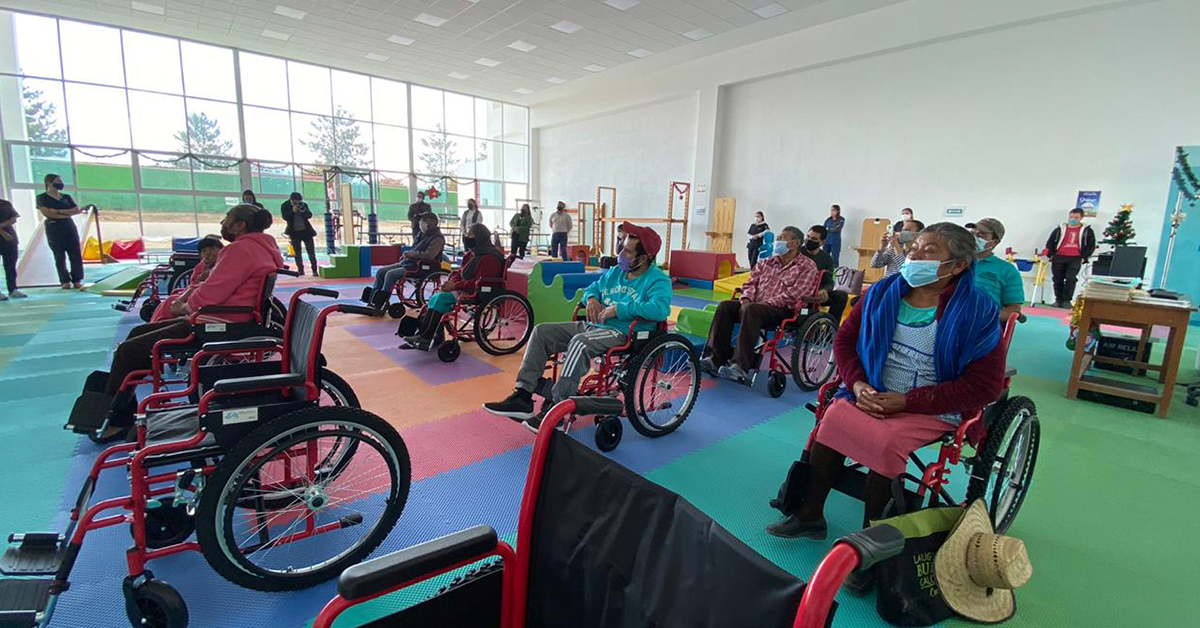 Fotografía de un salón donde se encuentran ocho personas sentadas en silla de ruedas, el piso es de colores y hay algunas sillas sin personas sentadas.