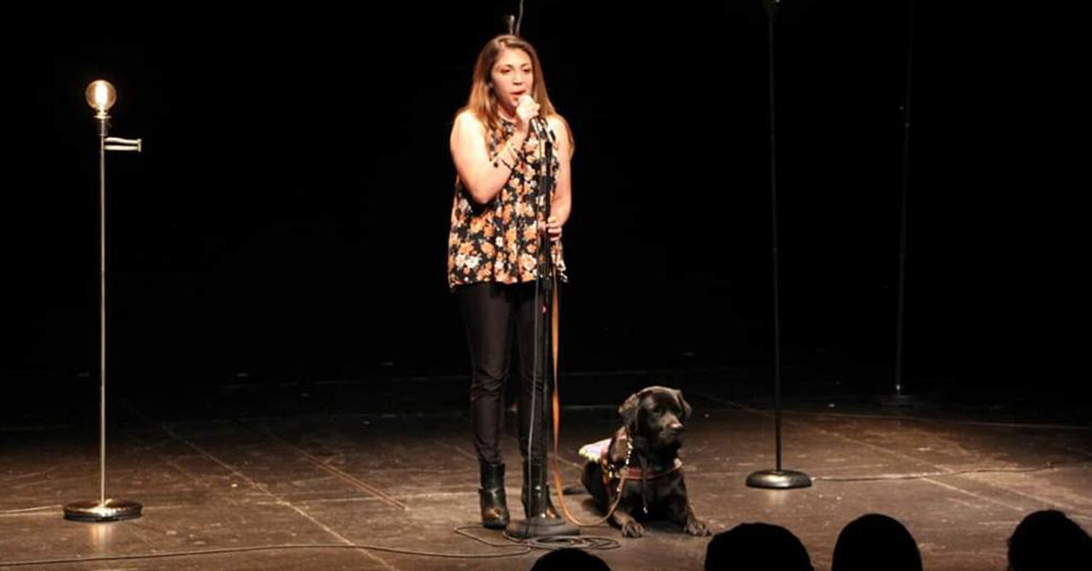 Fotografía de Erika de pie en un escenario, viste una blusa negra con un estampado de flores de colores y un pantalón negro. A su derecha está acostado un perro y frente a ella un micrófono.