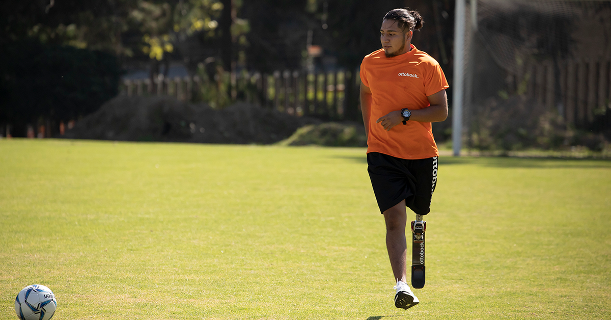 Fotografía de Gustavo en una cancha de futbol, está de frente corriendo hacia un balón que está frente a él. Lleva puesta una playera naranja y un short negro.