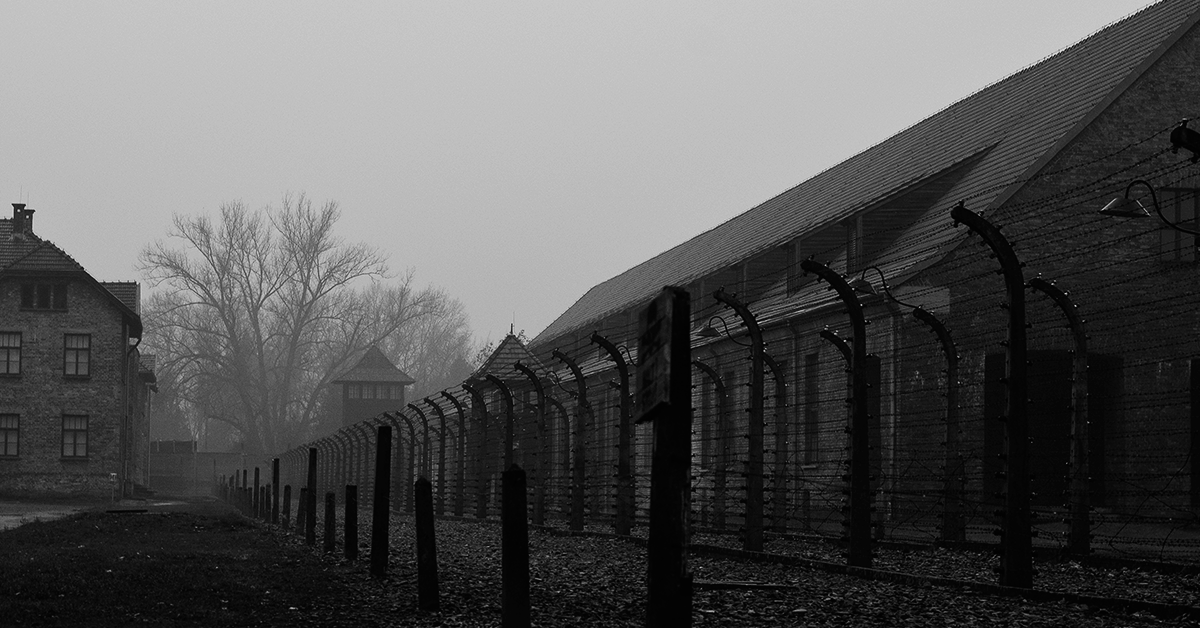 Fotografía en blanco y negro de un campo de concentración en Alemania, al frente una reja y al fondo un edificio, junto a él hay árboles.