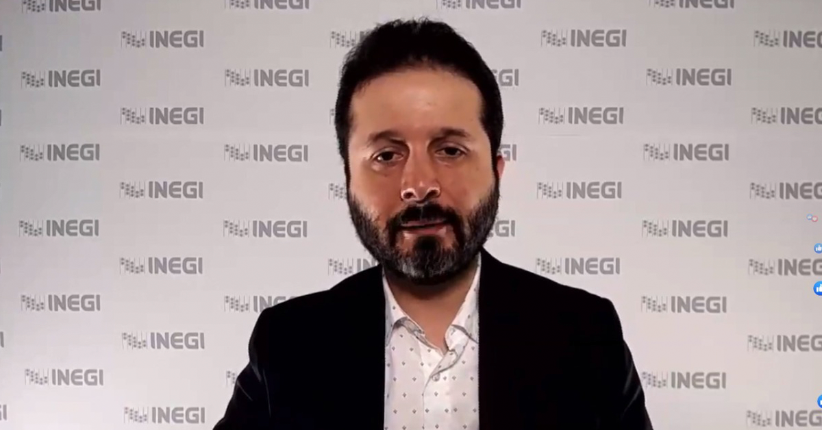 Fotografía del rostro de Edgar viendo directamente a la cámara mientras sonríe, detrás de él hay un cartel con el logotipo del INEGI impreso en color gris.