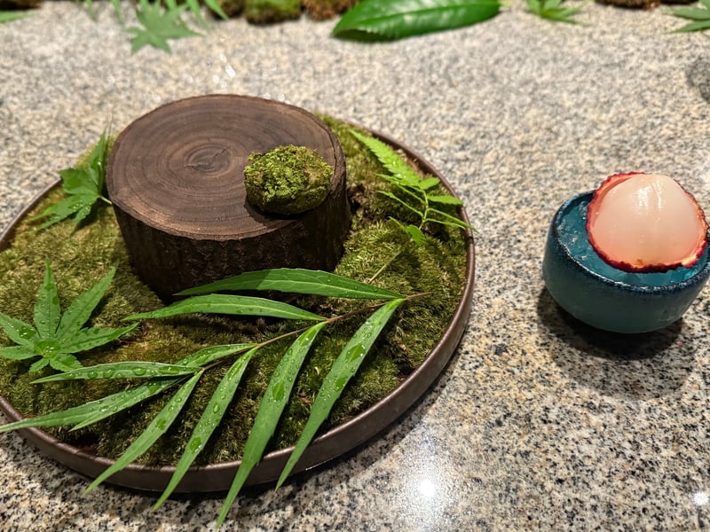苔を敷いた盆の切り株の上に、抹茶をまぶした一口菓子。横にライチの器