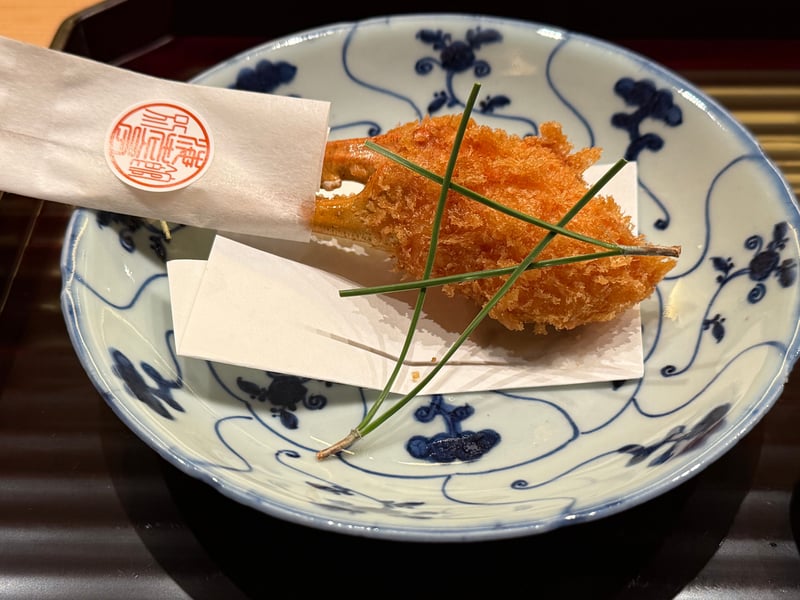 染付の皿に蟹の爪のフライ。松葉を交差させ、店の朱印の紙を敷く