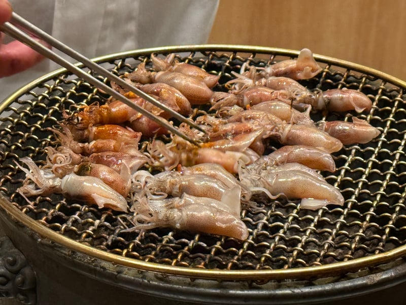 丸い炭火の焼き台に並べたホタルイカを、箸で返すところ