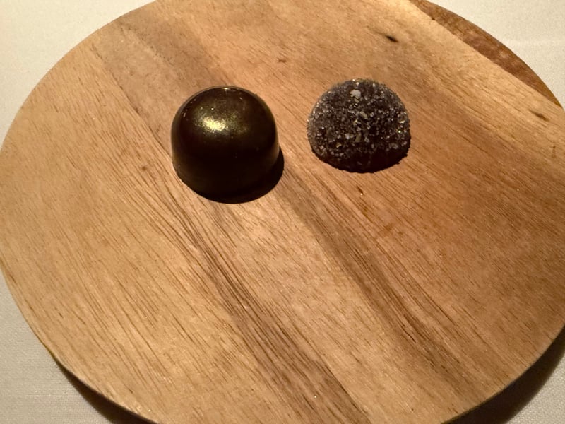 木の丸皿に小菓子が2つ。艶のある黒いドームと、砂糖をまとったもの