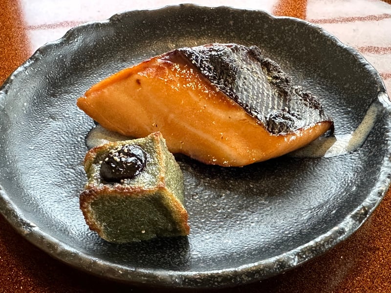 黒い皿に盛られた皮付きの焼き魚と緑の角切り