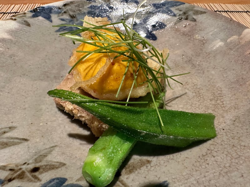 鰻の蒲焼きフライの上に橙色の卵黄の天ぷら。オクラと細い青い薬味が添えられている