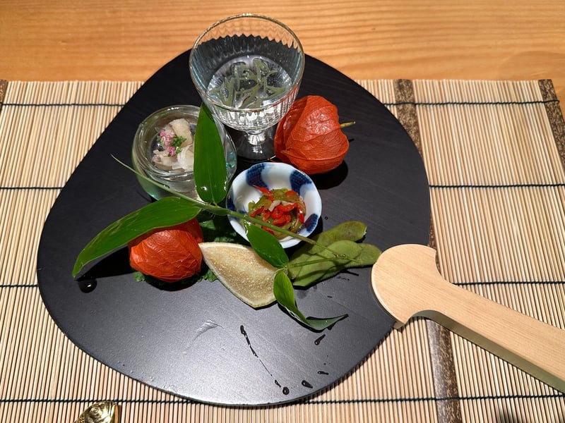 扇形の黒い皿に並ぶ八寸。ガラスの器に和え物、小鉢に万願寺とうがらし、枝豆と鬼灯が添えられている