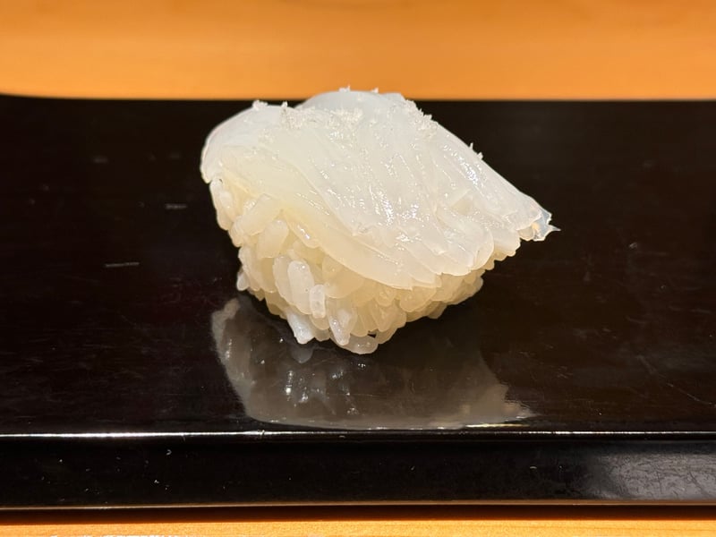 細かく包丁を入れたアオリイカの握り。黒い塗りの台に一貫