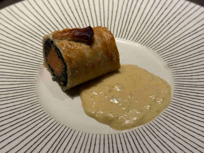 パイ生地で巻いて焼いた虹鱒。断面に橙色の身と緑の葉が層になって見え、隣にマスタードソース