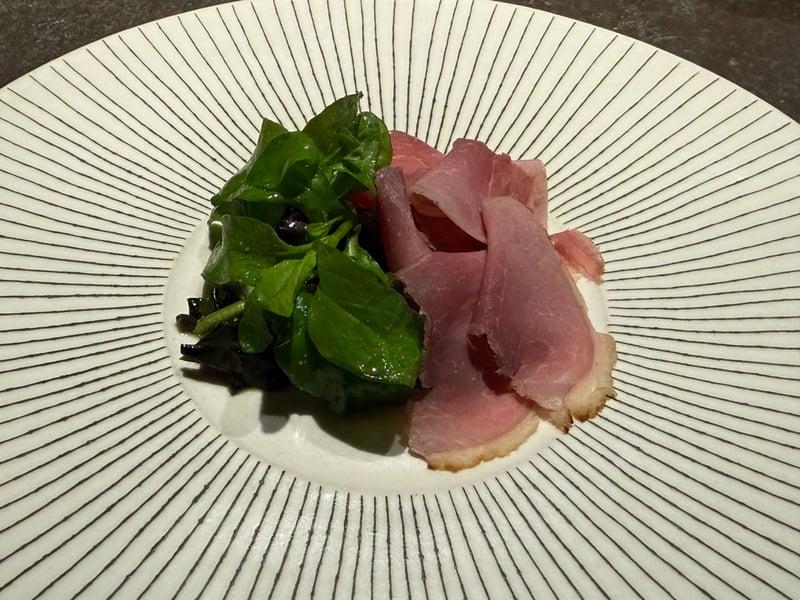 白いソースの上に薄切りの猪肉。横に緑の葉と黒紫の葉