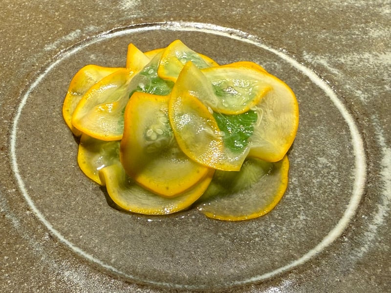 灰色の皿に、黄色い縁の薄切りズッキーニが重ねてある。下に緑のピューレ