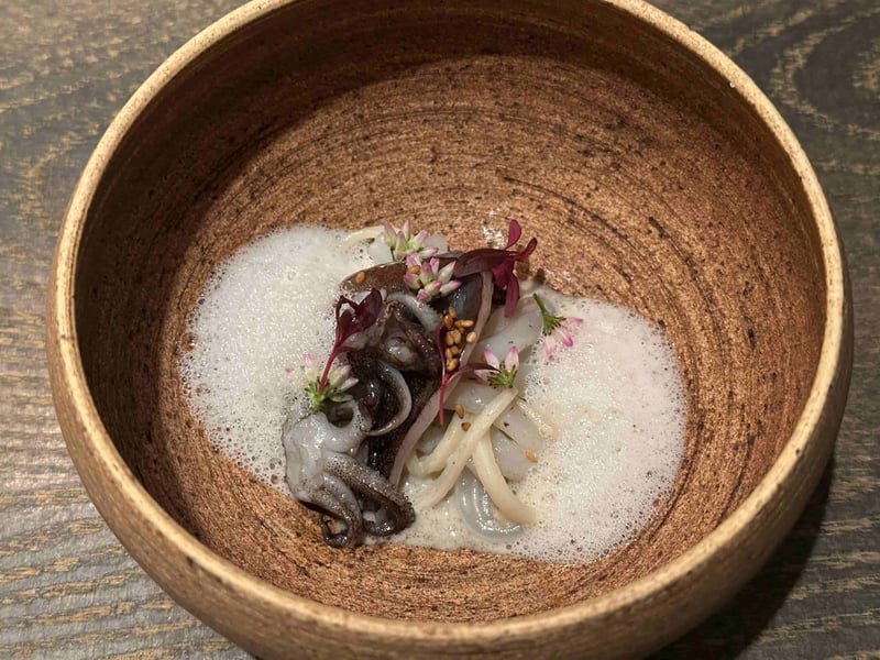 茶色の鉢に、白い泡をまとったアオリイカとマコモダケ。金胡麻と紫の花