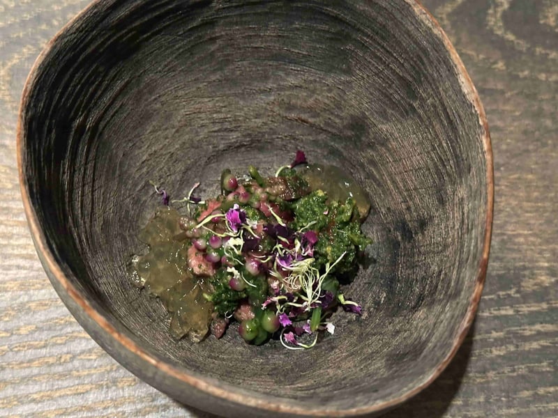 黒い鉢に月ノ輪熊の赤身。緑の山菜のソースと紫の花、透明なジュレ