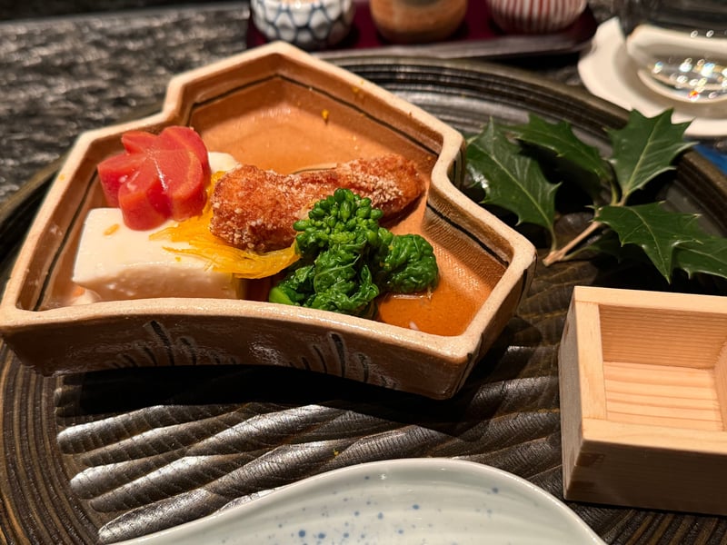 家形の器に白い豆腐と梅の形に抜いた赤いもの、黄色い細切り、衣をつけて揚げた小さなものが二つ茶色いソースに浸り、脇に菜の花
