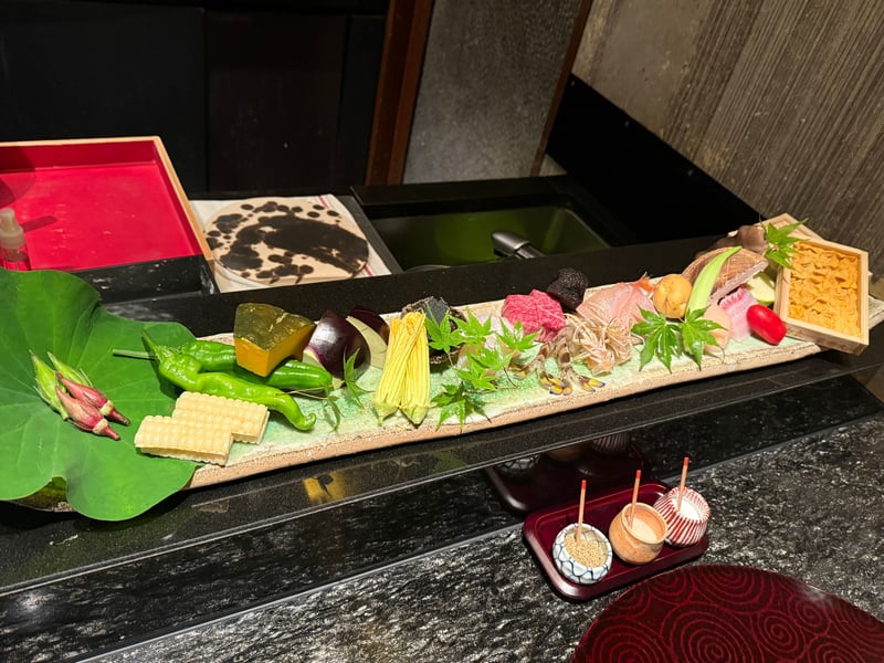 長皿に並んだこの日の食材。蓮の葉の上のオクラのつぼみ、白いとうもろこし、唐辛子、かぼちゃ、賀茂茄子、かぼちゃの花、近江牛と黒トリュフ、生きた車海老、じゃがいも、魚の身、木箱の雲丹