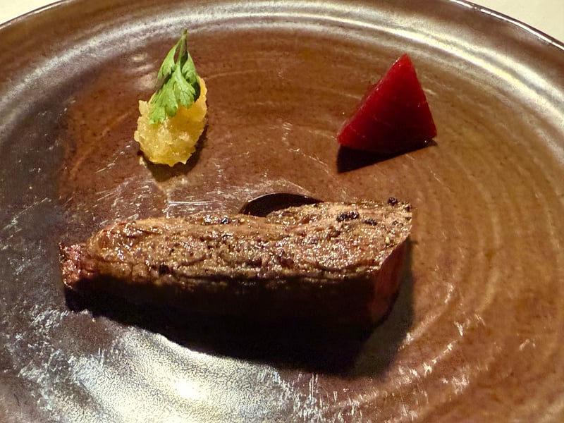 茶色い皿に蝦夷鹿の炭火焼き。リンゴのコンポートとセルフィーユ、赤いくさび形が添えてある