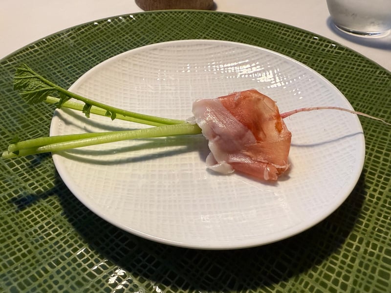 緑の器に置いた白い皿。葉と細い根を残した小さな根菜に生ハムを巻いてある