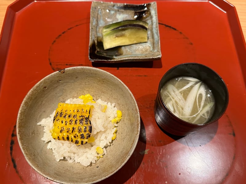 取り分けたとうもろこしご飯の椀。焼いたとうもろこしがひと切れのり、茄子の漬物と椀が並ぶ