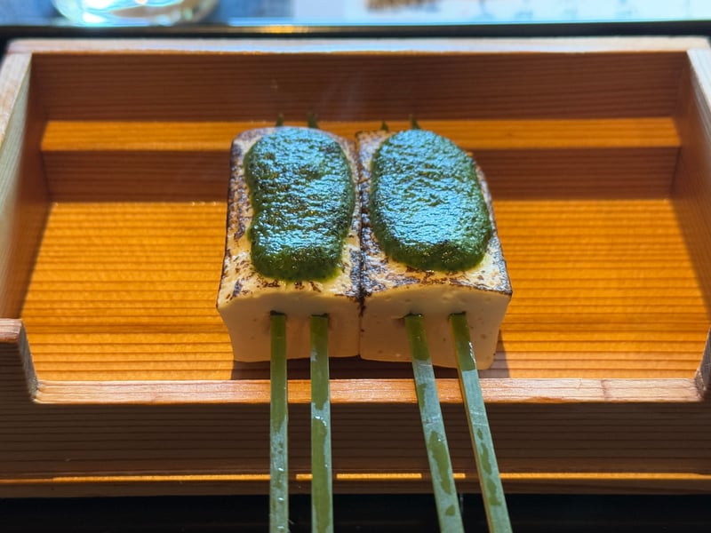 木箱に載せた二本の豆富の田楽。緑の木の芽味噌を塗り、青竹の串を刺してある