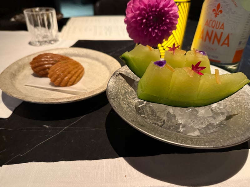 氷を敷いた皿に櫛形のメロン、食用花を散らしてある。左の皿に黄人参のマドレーヌが二つ