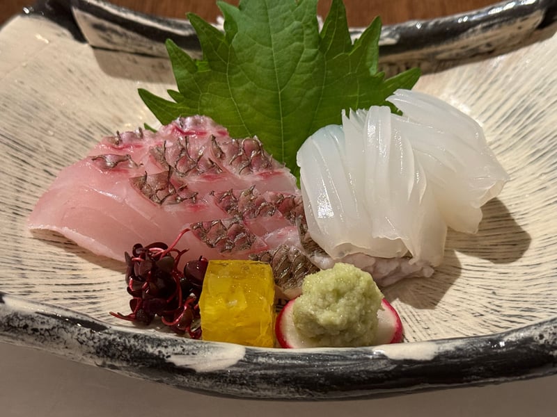 黒縁の皿に、松皮造りの真鯛と白い水烏賊。大葉とわさび