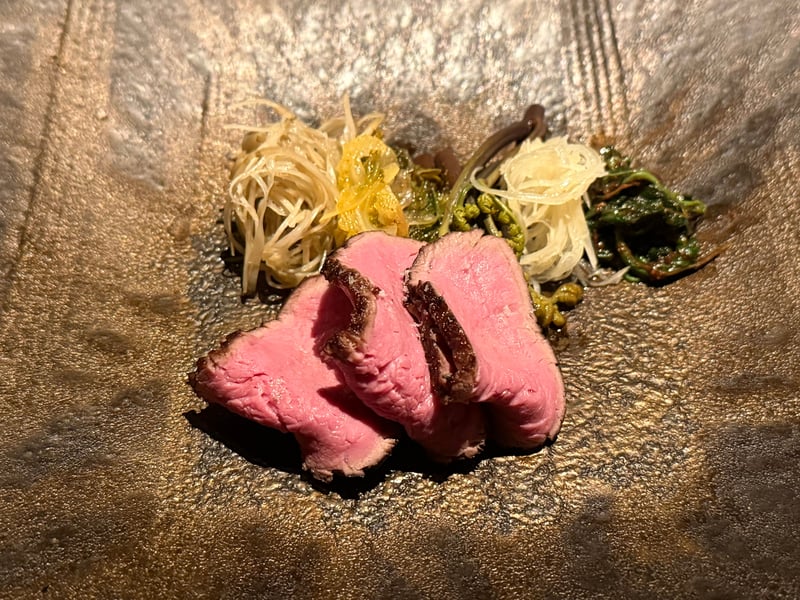 石肌の皿にのった短角牛の薄切り三枚。奥に牛蒡と山菜が添えられている